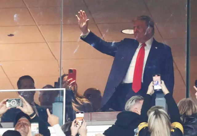 Donald Trump waves at at crowd from behind a glass window, there are several mobile phones pointed at him.