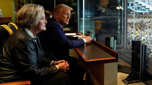 Donald Trump sits next to a woman in a private booth at a stadium American football game. He is seen looking through the glass of the private booth, and the woman sitting next to him is older and wearing a black leather jacket.