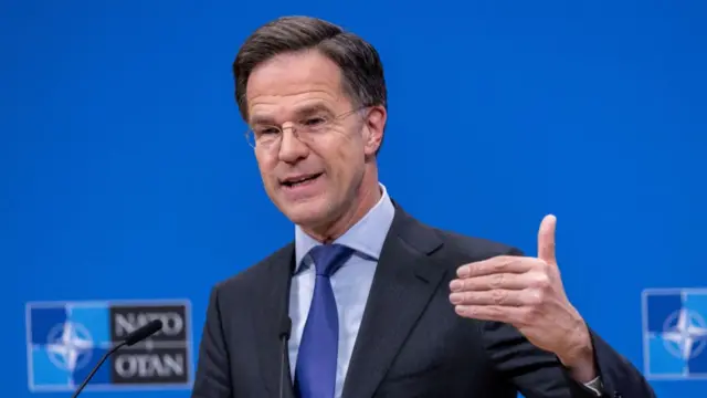 Mark Rutte in dark suit with blur tie with hand partly up in the air. He is speaking into a microphone with a blue wall behind him.