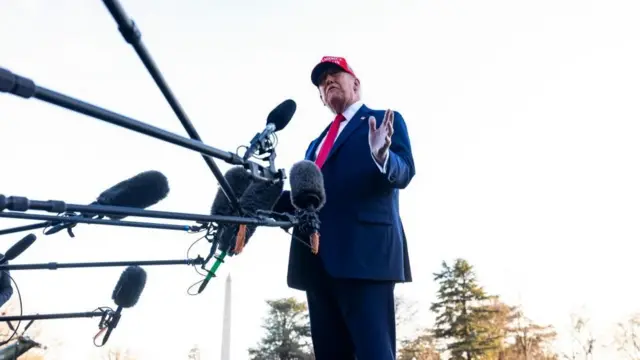 Trump wears a red tie and MAGA hat and answers questions outside with multiple microphones directed at him.