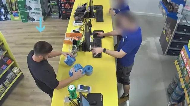 Kyle Clifford wearing a black T-shirt inside a Toolstation store. He has been handed three pieces of rope from a member of staff the other side of a yellow counter