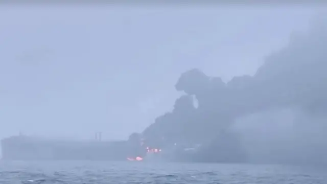 A grey scene on the sea, where a boat appears to be on fire. It's hard to make out, but there are orange flames and large amounts of grey smoke rising from the wreckage