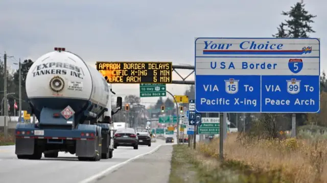 A tank drives down a highway with a sign on the left reading 'USA border' and a sign above warning of delays at the border