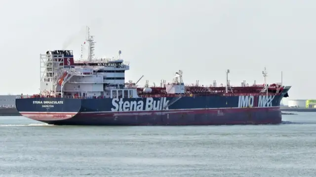 The Stena Immaculate, which has those words written on the rear of the ship in white, sits at sea. It is a red and white, long ship, with at one end a white structure with multiple levels