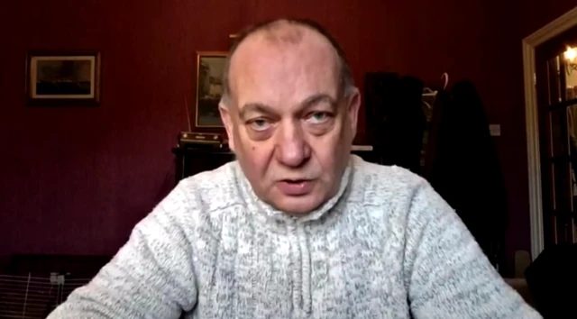 Headshot of Mcfarlane wearing a grey zipped up knit jumper. He sits in a room with red walls and paintings behind him.