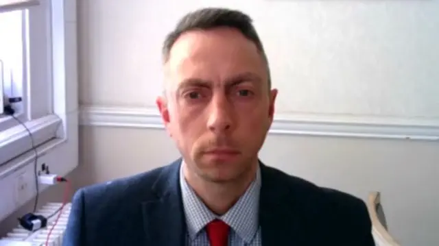 Headshot as he sits looking into the camera with a blank wall behind him and a window in the background to one side. He wears a suit jacket, blue check shirt and red tie