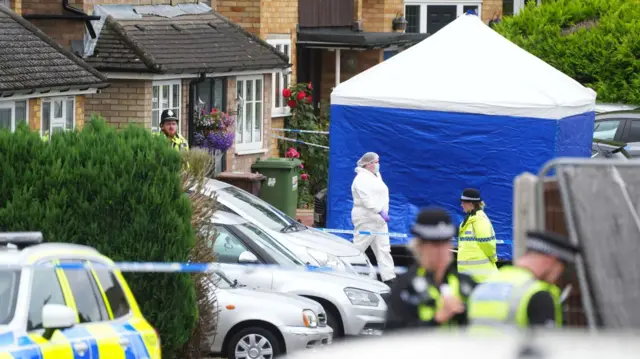 Police and a forensics officer outside a house in a residential road. The house is taped off and has a blue and white police tent outside it.