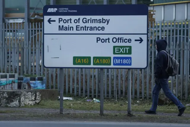 A person in jeans and a hoodie walking past a road sign. It points right to the port office, and left to the port of Grimsby main entrance