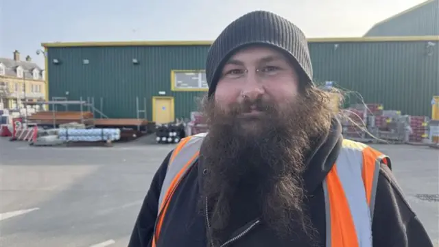 A bearded man smiling at the camera. He has a piercing in his nostril and between his eyebrows. He has a beanie on, a black hoodie and a high vis and stands in a card part