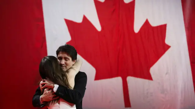 Trudeau faces the camera and hugs his daughter - the Canadian flag is in the background