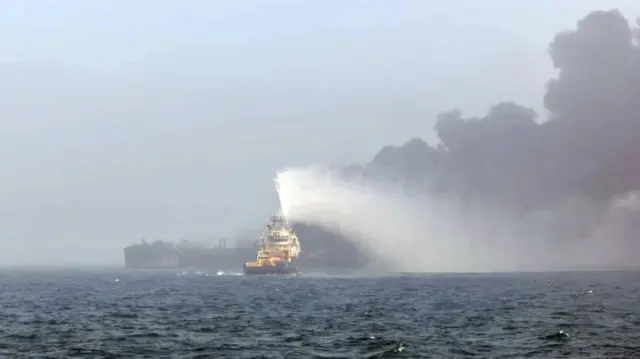 A large boat seen from side on is burnt out and black. A smaller ship in front of it blasts water at the burning boat, which is surrounded by black smoke