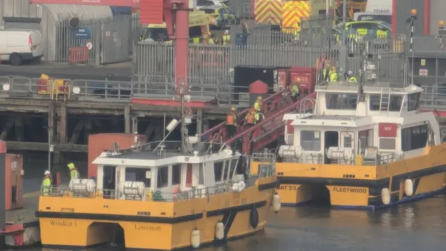 a close up shows crew members in high-vis vests arriving on the shore line. two rescue vessels can be seen to the side.