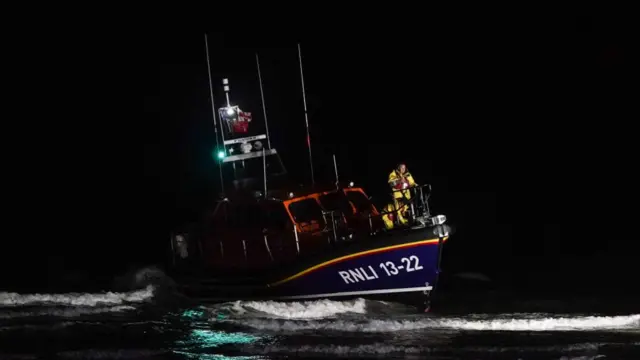 An RNLI boat in the night moves along in the water. It has a person at the bow wearing a yellow rubber suit and the words RNLI 1322 are on the front in white letters. The rest of the boat is blue and orange