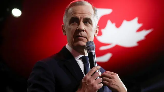 Mark Carney stands in close up holds a microphone and has a Liberal logo in the background