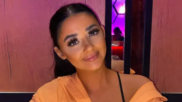 Louise Hunt with her black hair in a ponytail. She is wearing an orange shirt off her shoulder and is smiling while sitting in a restaurant.