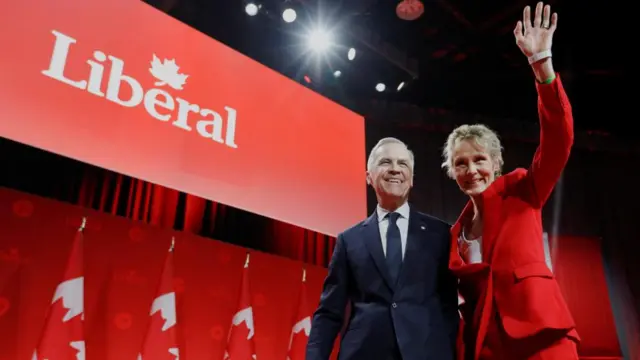 New party leader Mark Carney smiles and his wife Diana Fox, who is next to him, waves on stage after he won the leadership contest. A red banner that says 'Liberal' is in the backround.