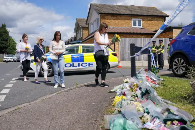 Four women lay flowers at the entrance to the road where the Hunt family live. Police officers watch on as they do so.