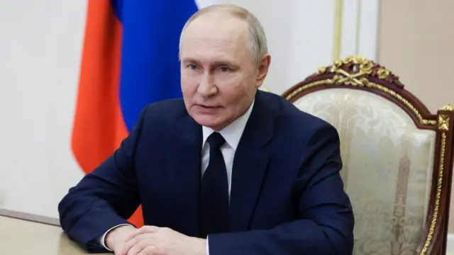 Vladimir Putin sits in a chair wearing a suit with a Russian flag in the background.