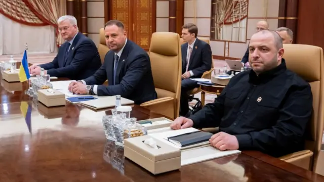 The three Ukrainian delegates sit at a wooden table. In front of them are notebooks, refreshments and small Ukrainian flags.