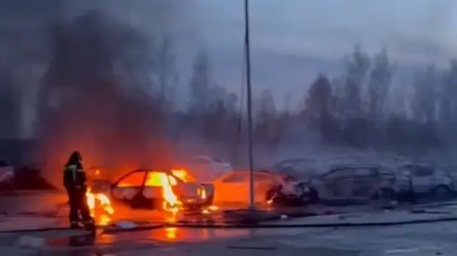 A firefighter putting out a car on fire in a car park with dark smoke coming out of it