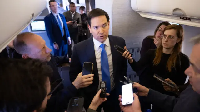 Marco Rubio, wears a suit, surrounded by reporters on a plane.