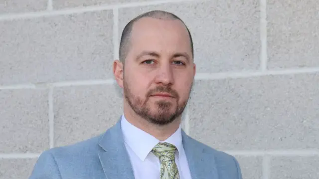 Nick Gardner wearing a light grey suit, yellow tie and white shirt. He looks serious in his expression