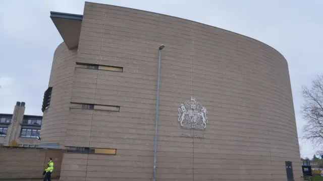 The beige Cambridge Crown Court building, which has a silver crest on it and a person wearing a hi-vis outside. It is a large building of multiple storeys.