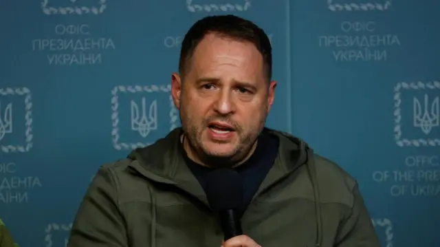 Andriy Yermak wears a zip up dark green hoodie with a black top holding a microphone.