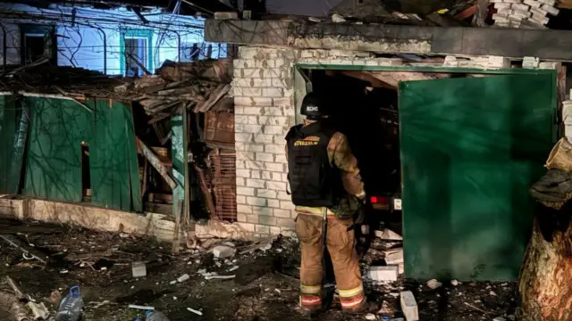 An emergency service worker stands in front of a ruined building. A fence and door has been knocked down, and there is some rubble at the mans feet