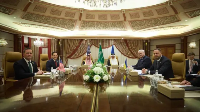 Delegates for Ukraine, US and Saudi Arabia seated round a table. Each have their country's flag positioned in front of them.