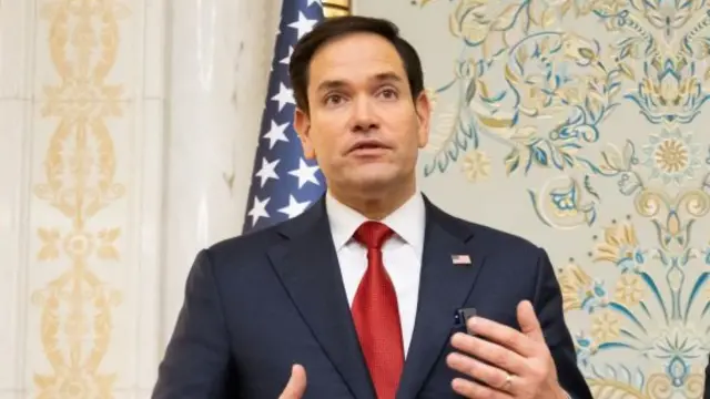 Marco Rubio in a suit and red tie, stood in front of a patterned wallpaper and US flag.