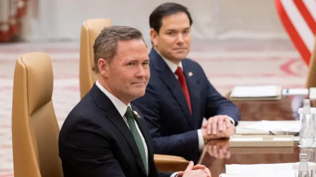 The two US delegates sit at a wooden table. In front of them are notebooks and refreshments.