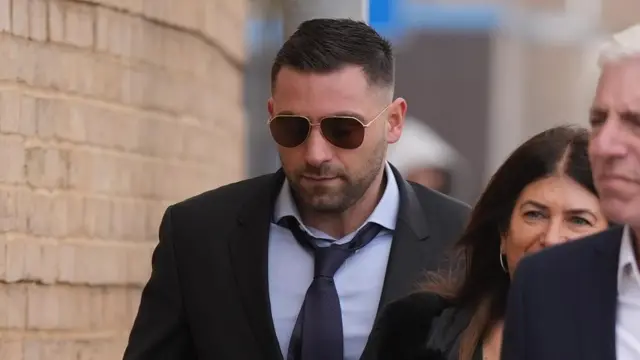 Alex Klein wearing a dark suit, light blue shirt and navy tie. He has dark hair, light stubble and is wearing sunglasses