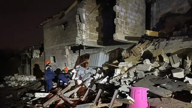 A building that's side has fallen down. There is rubble everywhere. Two emergency workers look up at the damage