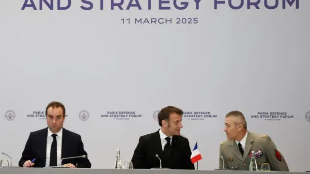 Macron sits in the middle of a desk with a small French flag in front of him. To one side is a man in a suit, and the other a military chief who speaks with Macron