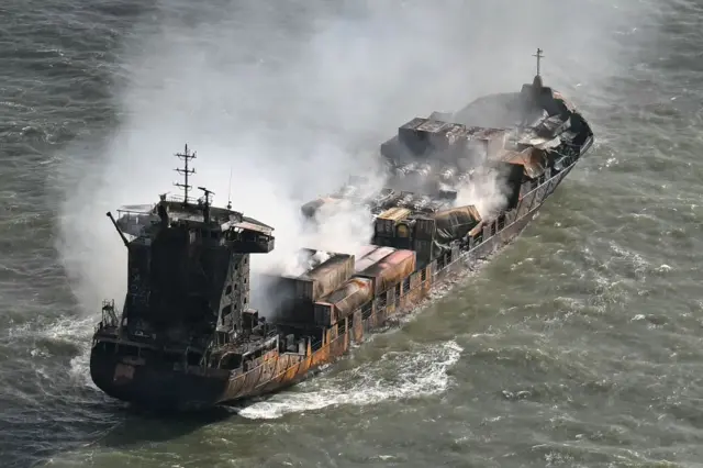 A burnt out cargo ship with its cargo containers brown and smoke pouring off the top of the ship. The water around it is a dark greyish green colour