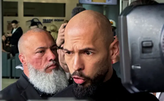 Andrew Tate, who has a shaved head and black beard, speaks to reporters. There is a black TV camera to the right and a security guard to the left.