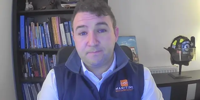 Matthew Shanck with short brown curly hair and fleece vest with the Maritime Search and Rescue Council logo sitting in an office with a bookshelf and a fire rescue helmet.