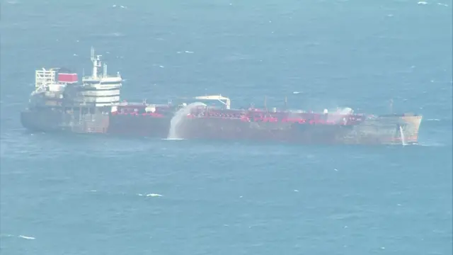 A tanker is seen with water pouring out of it in the sea. It is red.