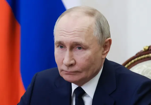 Putin in a navy suit and tie and a straight face sat in front of a Russian flag