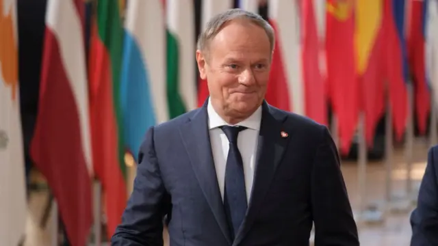 Tusk walking along in front of multiple flags. He wears a suit and smiles