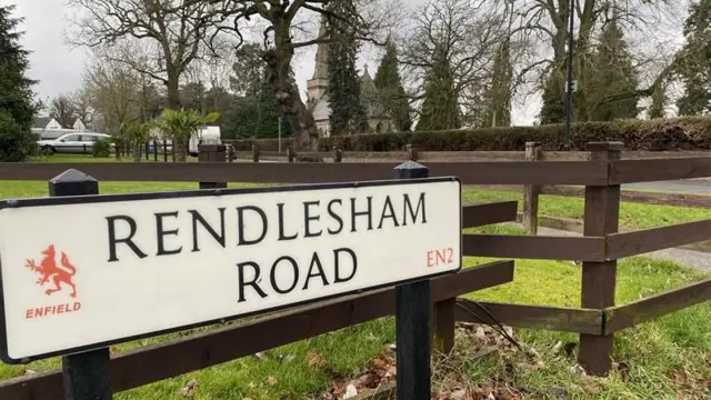 A sign for Rendlesham Road, which is attached to a brown fence. It is at the entrance to a cemetery, which is surrounded by bushes and trees.