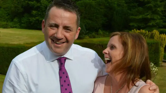 John Hunt wearing a white shirt and pink tie with black polka dots. He is smiling and has is arm around Carol Hunt, who is wearing a dress and laughing while looking at him.