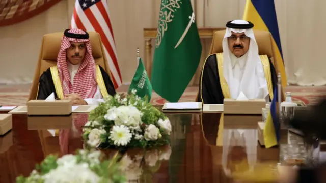 The two officials sit at the head of the table in front of notebooks and a small Saudi flag