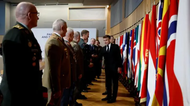 Macron in front of a line of military men, as he shakes one of their hands. Behind him there is a line of flags rom different country's