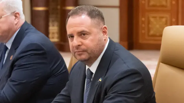 Yermak sits at a table in a beige chair and looks to his side. He wears a suit with a small badge of the Ukrainian flag