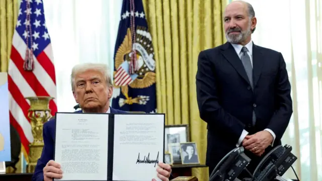 Donald Trump holds an executive order about tariffs increase in February, flanked by Commerce Secretary Howard Lutnick in the Oval Office. Both men are wearing suits, Trump is sitting down while Lutnick is standing.