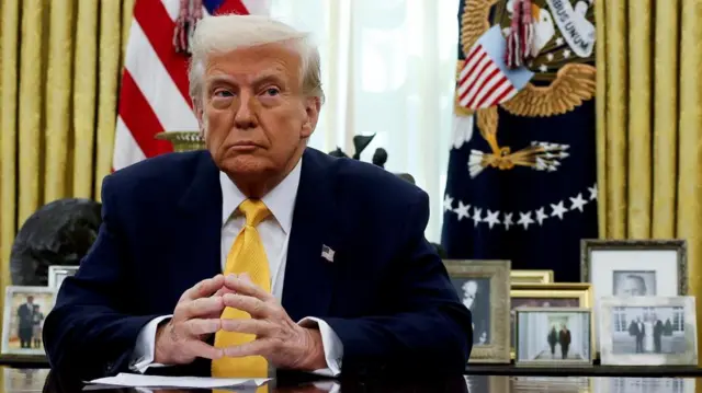 Donald Trump looks to his left as he's sat in the Oval Office wearing a suit and yellow tie.