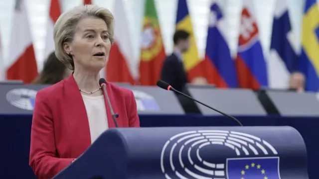 European Commission President Ursula von der Leyen speaking at the European Parliament. She has a lectern with a mic in front of her and flags of various countries in the background.