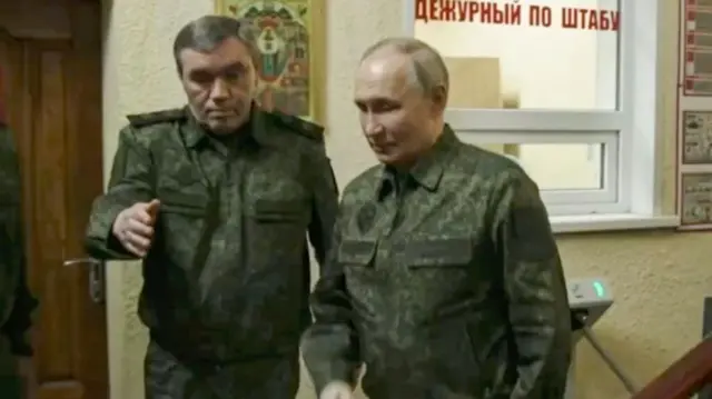 Vladimir Putin in military fatigues walks beside a military officer.